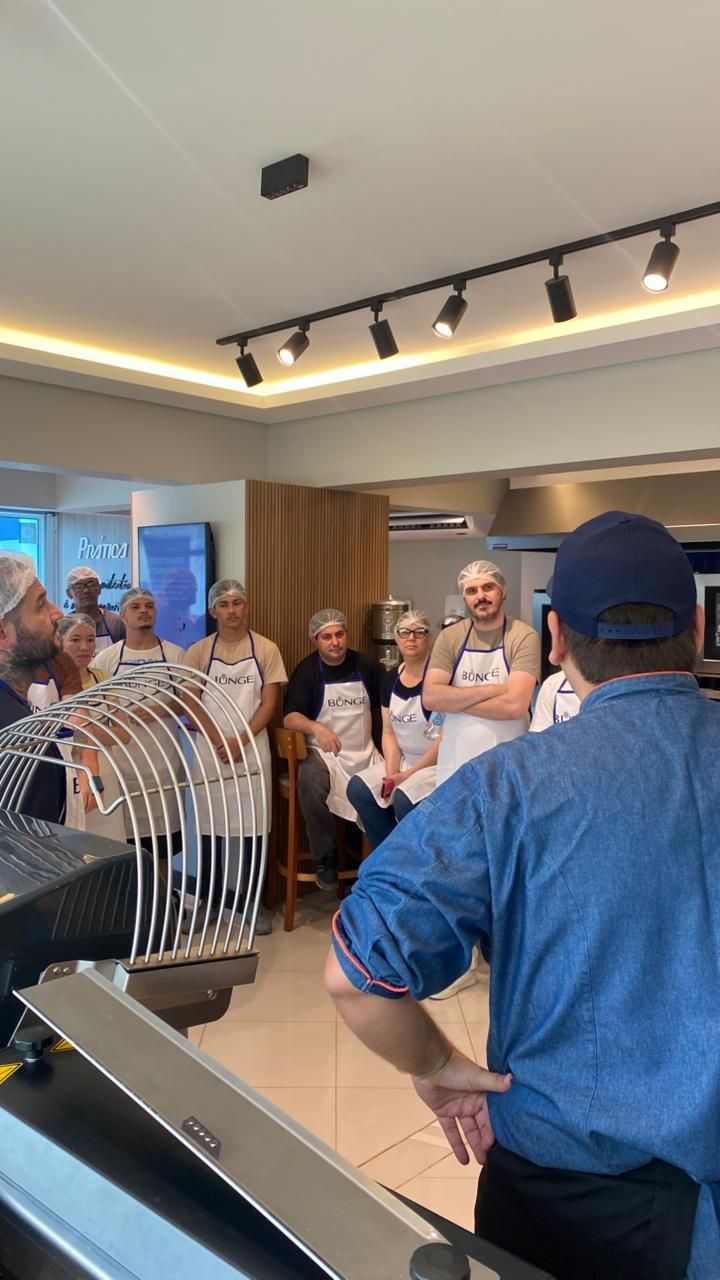 Um grupo de pessoas usando aventais e toucas de cabelo em uma cozinha ouve alguém dando instruções.