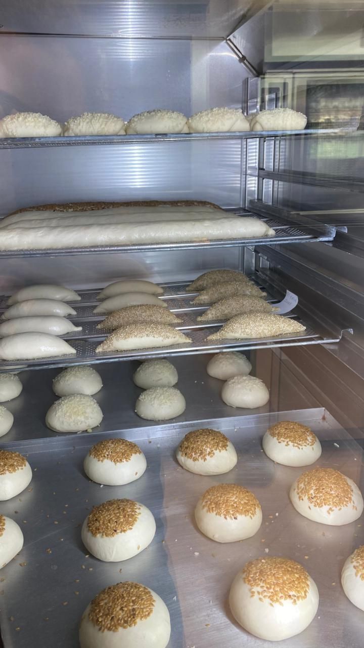 Massa de pão em forno industrial, de vários formatos, algumas com sementes de gergelim, em diversas prateleiras.