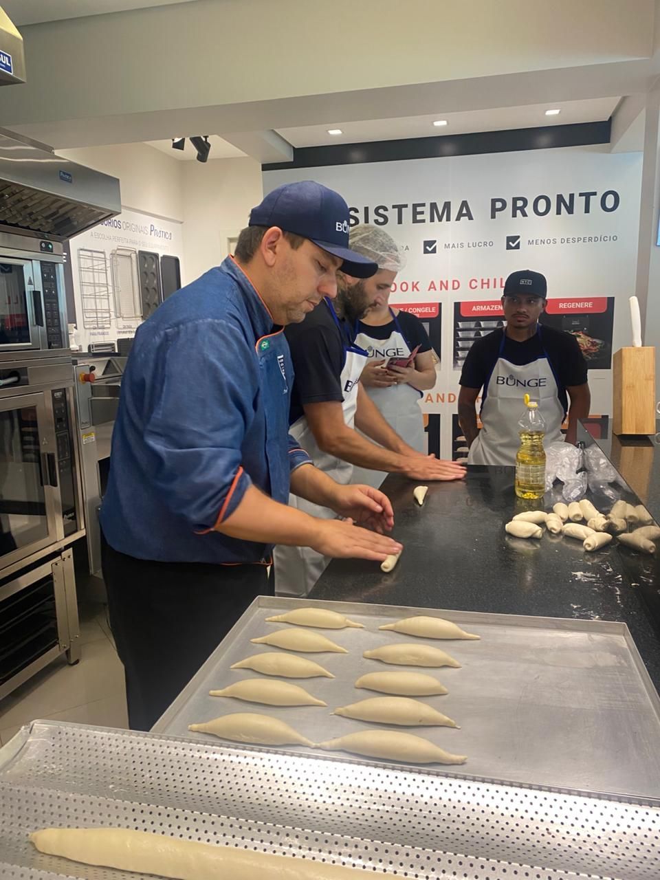 Pessoas moldando massa em uma bancada de cozinha; um padeiro está usando um casaco e um boné de chef azuis.