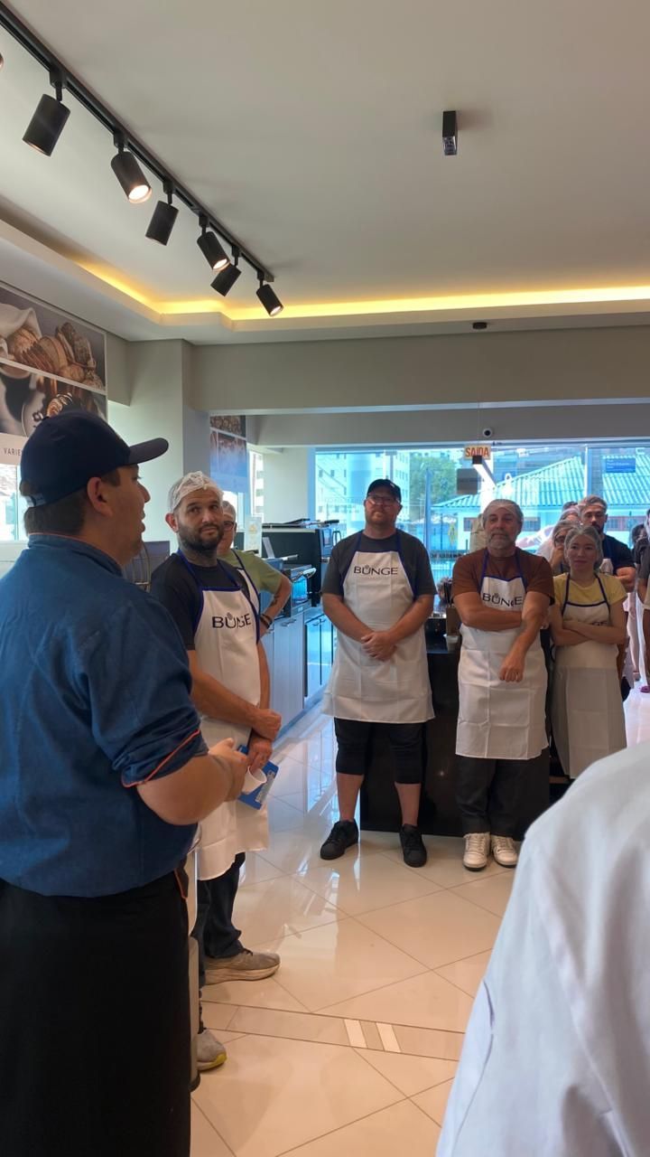 Grupo de pessoas de avental e chapéu, ouvindo alguém falar. Dentro de um café.