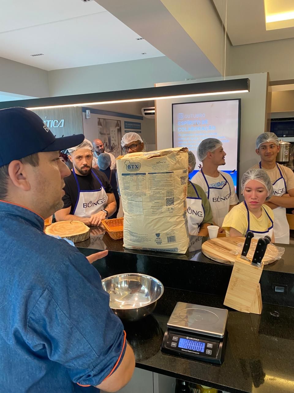 Chef demonstrando como fazer pão para um grupo em uma padaria. Ingredientes e utensílios sobre a bancada.