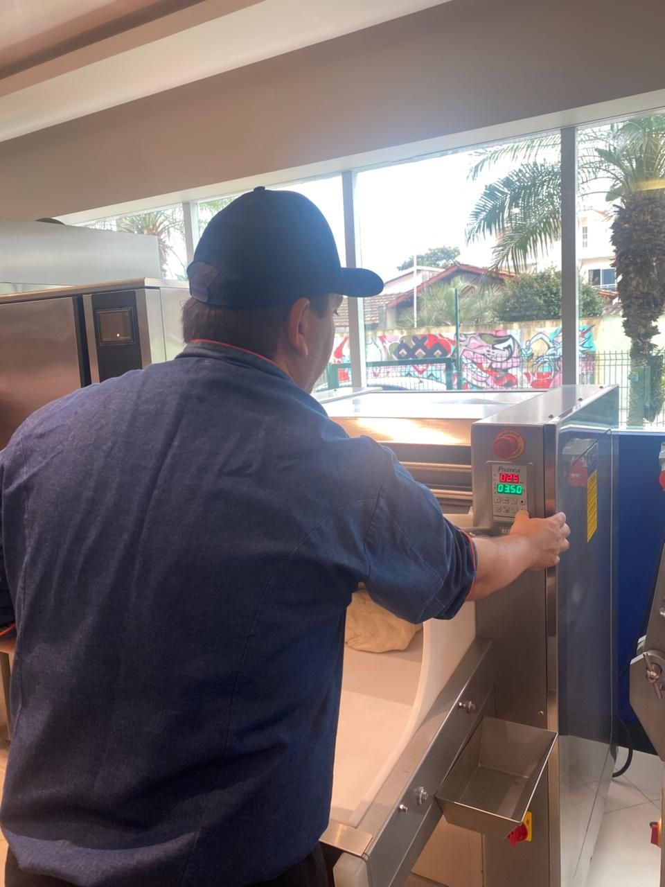 Uma pessoa vestindo camisa azul e boné de beisebol opera um forno industrial próximo a uma janela.