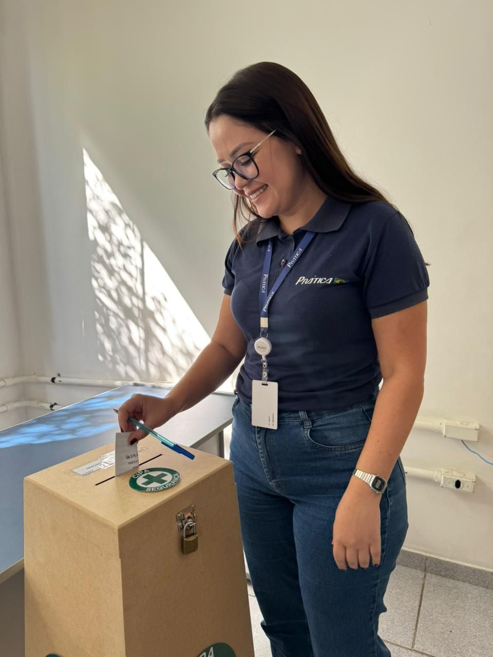 Mulher de camisa azul depositando um voto em uma urna de madeira. Ela está sorrindo. Fundo de parede branca.