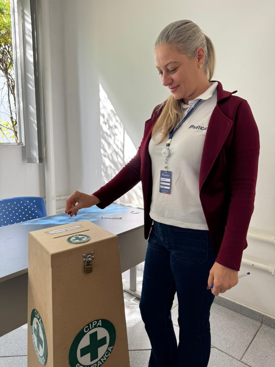 Mulher votando e colocando um papel em uma urna de papelão com um símbolo de segurança verde. Em ambiente interno, em pé perto de uma mesa.