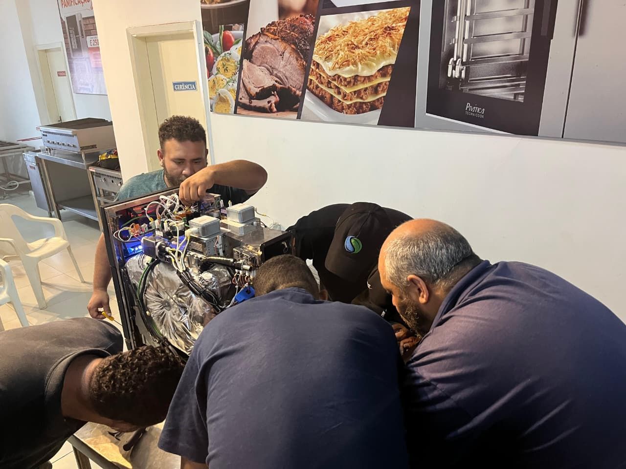 Quatro homens, incluindo um homem de camisa verde, consertam máquinas em uma cozinha.
