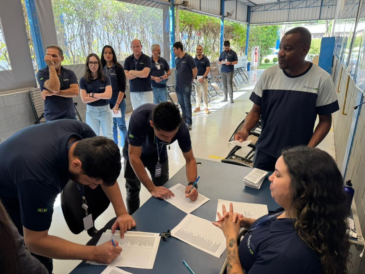 Pessoas assinando documentos em uma mesa, outras observando. Várias vestem camisas azul-escuras. Ao ar livre, em uma área coberta.