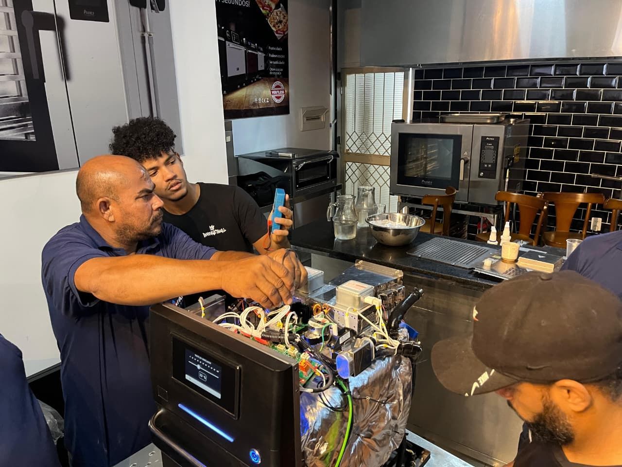 Pessoas trabalhando em uma máquina de café em uma cozinha comercial; uma delas segura um telefone.
