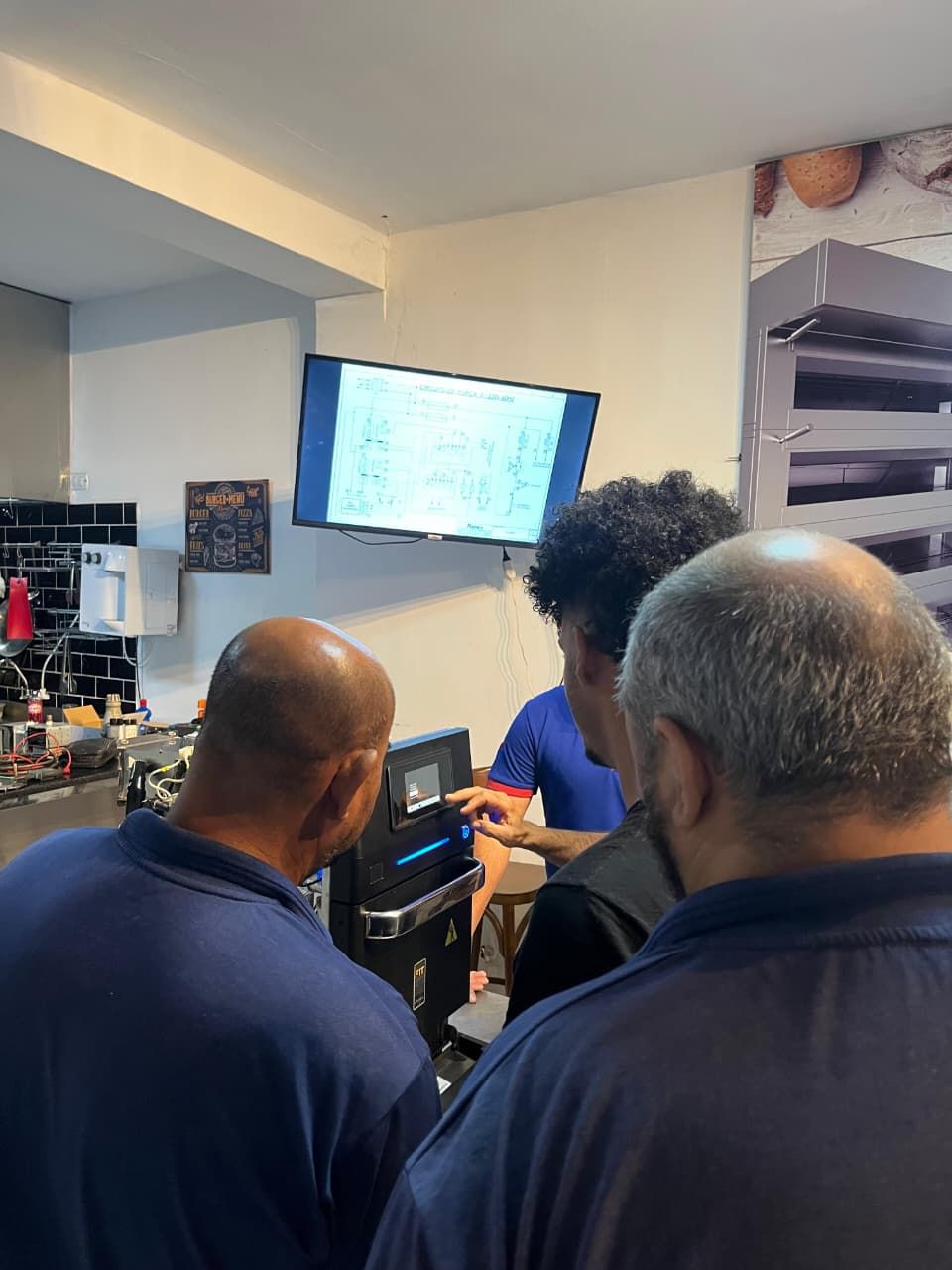 Três homens olhando para uma máquina, o homem no centro apontando. TV na parede. Interior de barbearia.