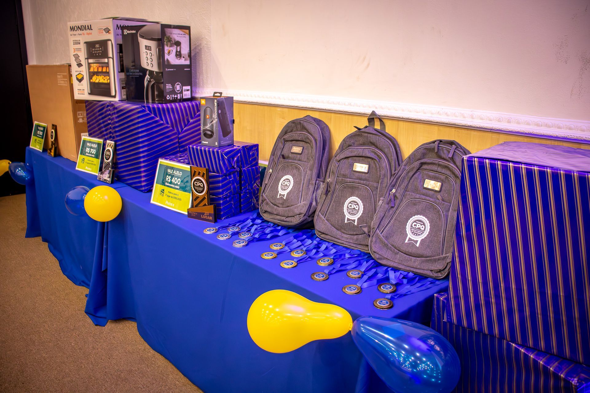 Prêmios em uma mesa coberta de azul e decorada com balões: presentes embrulhados, mochilas e medalhas.