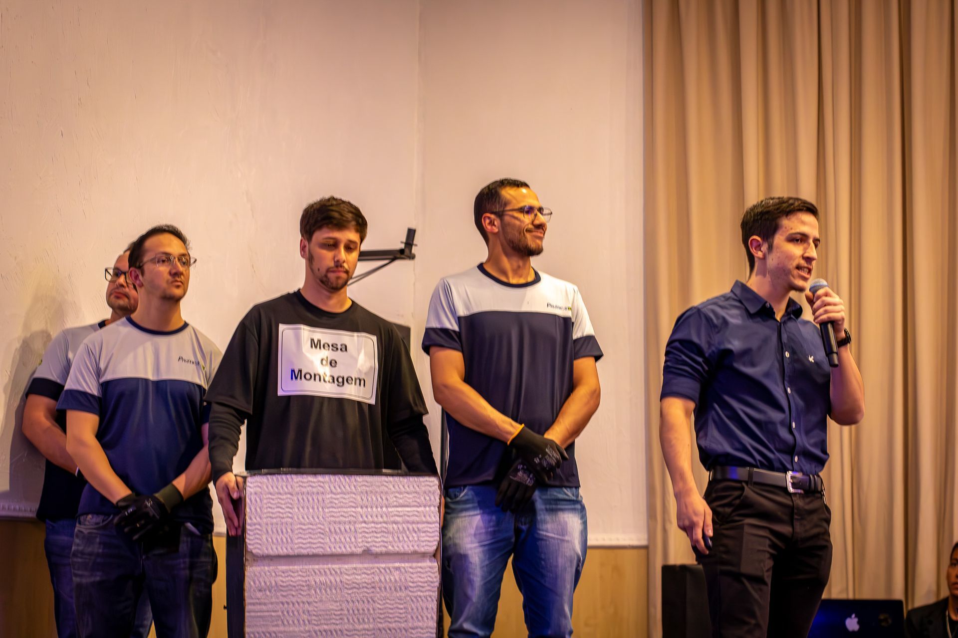 Cinco homens em pé em um palco. Um fala ao microfone; os outros observam. Os homens vestem roupas casuais. Uma estrutura branca e quadrada está à frente deles.