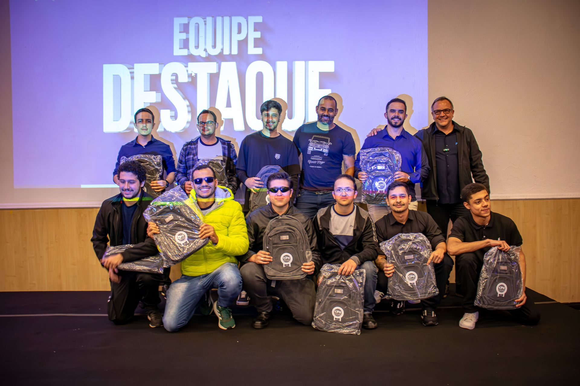 Um grupo de 13 homens, alguns ajoelhados, segurando mochilas, sorriem em frente ao cenário 