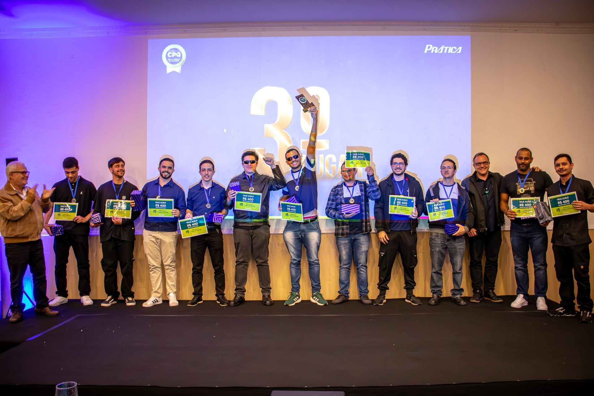 Um grupo de homens comemora em um palco, alguns segurando prêmios. Um telão exibe 