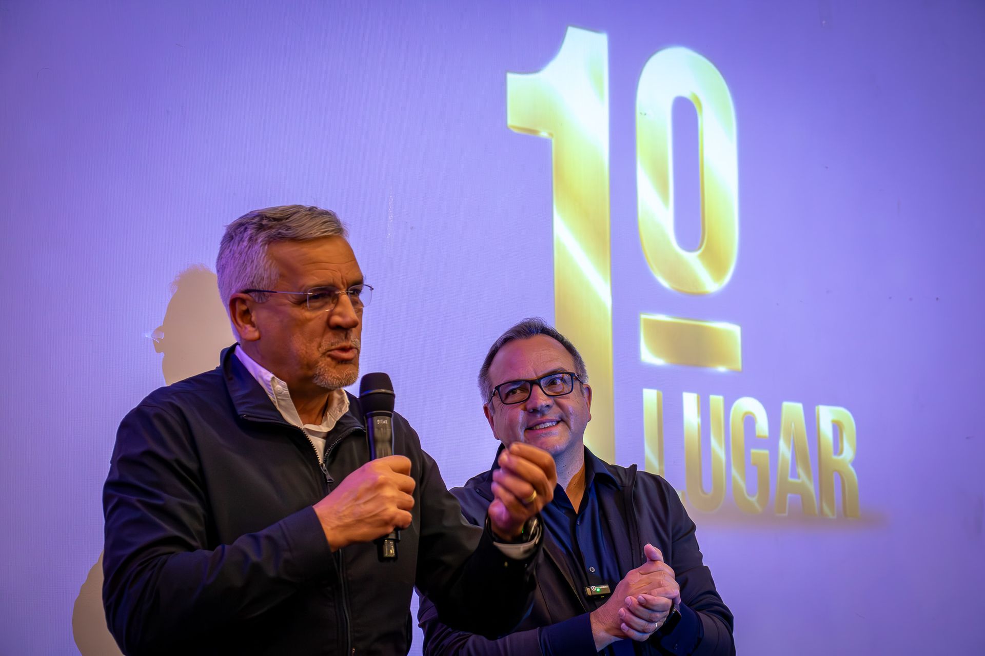 Dois homens estão em frente a uma tela onde se lê 
