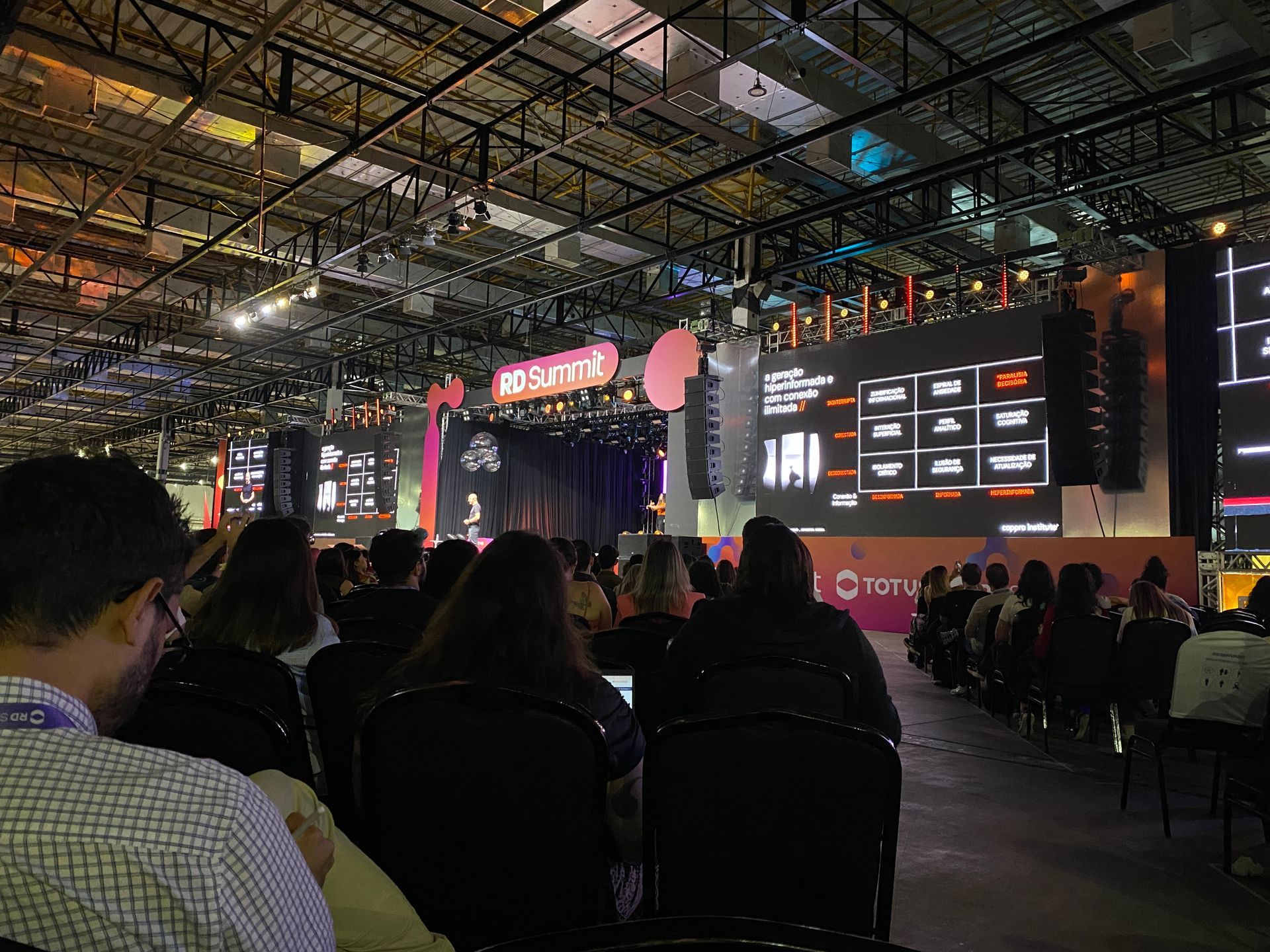 Público sentado, de frente para um palco com um apresentador, telões e o logotipo do 
