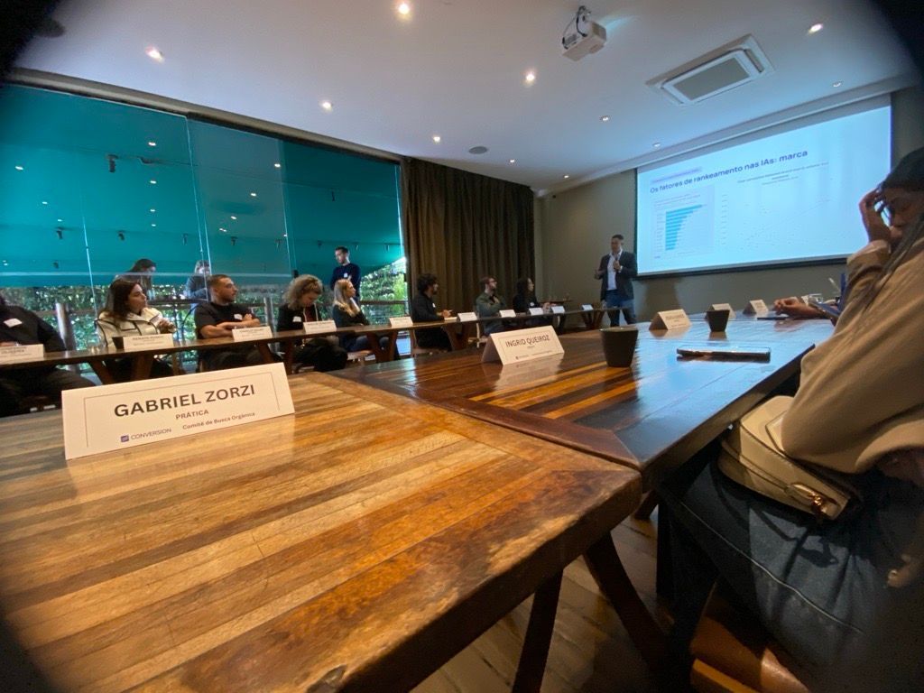 Uma sala de conferências com pessoas sentadas em uma longa mesa. Um apresentador está perto de uma tela com um gráfico.