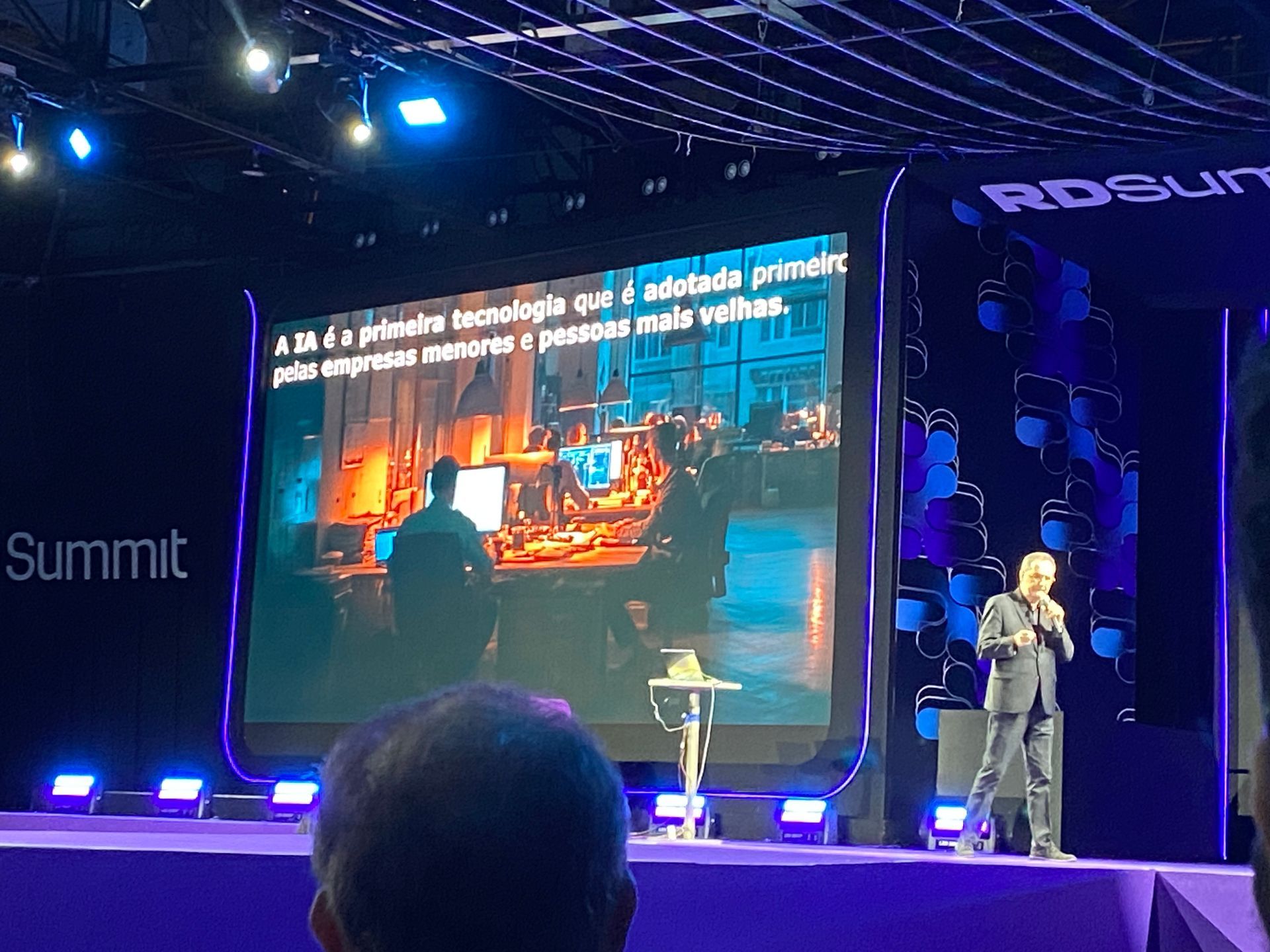 Homem discursando em um palco durante uma conferência; uma tela exibe uma imagem de pessoas e texto. Iluminação roxa.