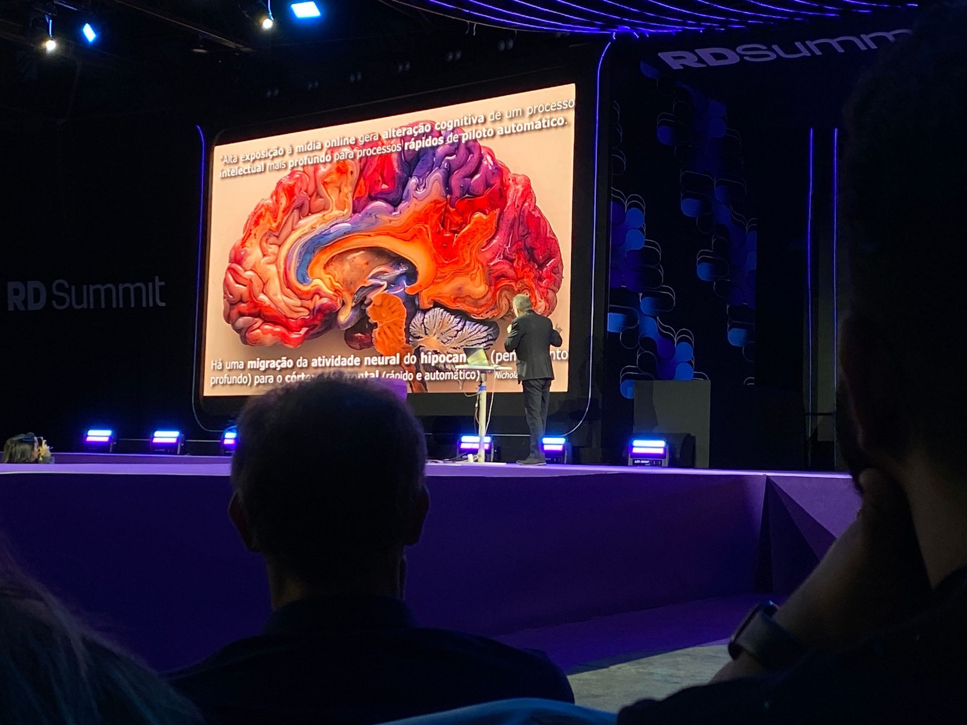 Homem apresentando exame de imagem cerebral no palco de uma conferência, visto da plateia.