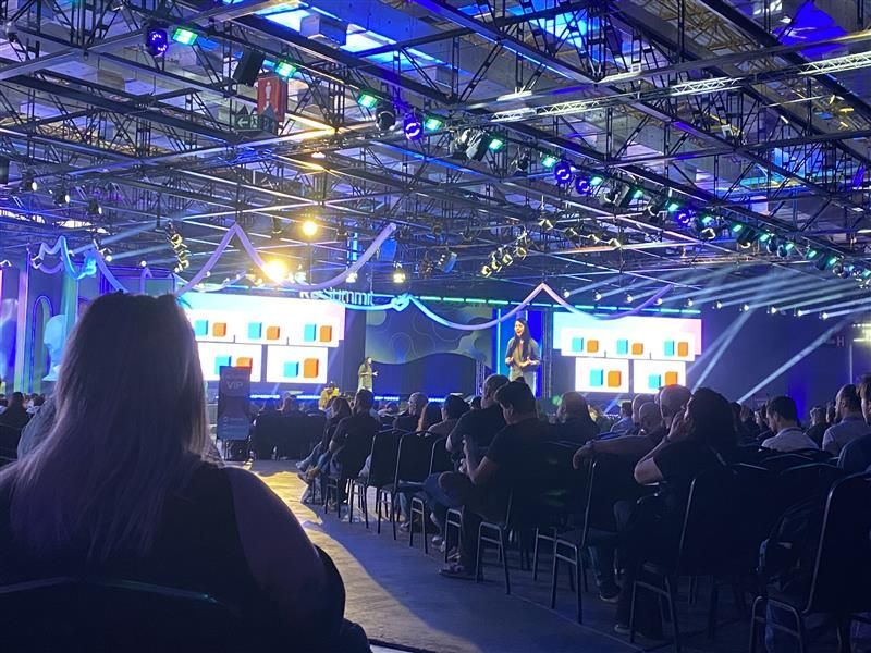 Público em uma conferência assistindo a uma apresentação no palco, intensamente iluminado por luzes azuis.