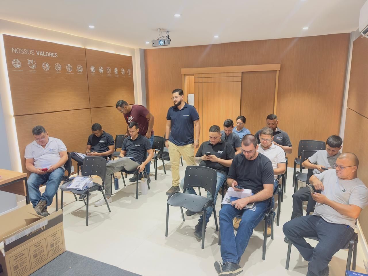 Um grupo de pessoas está sentado em cadeiras dispostas em uma sala de aula, algumas olhando para seus celulares.