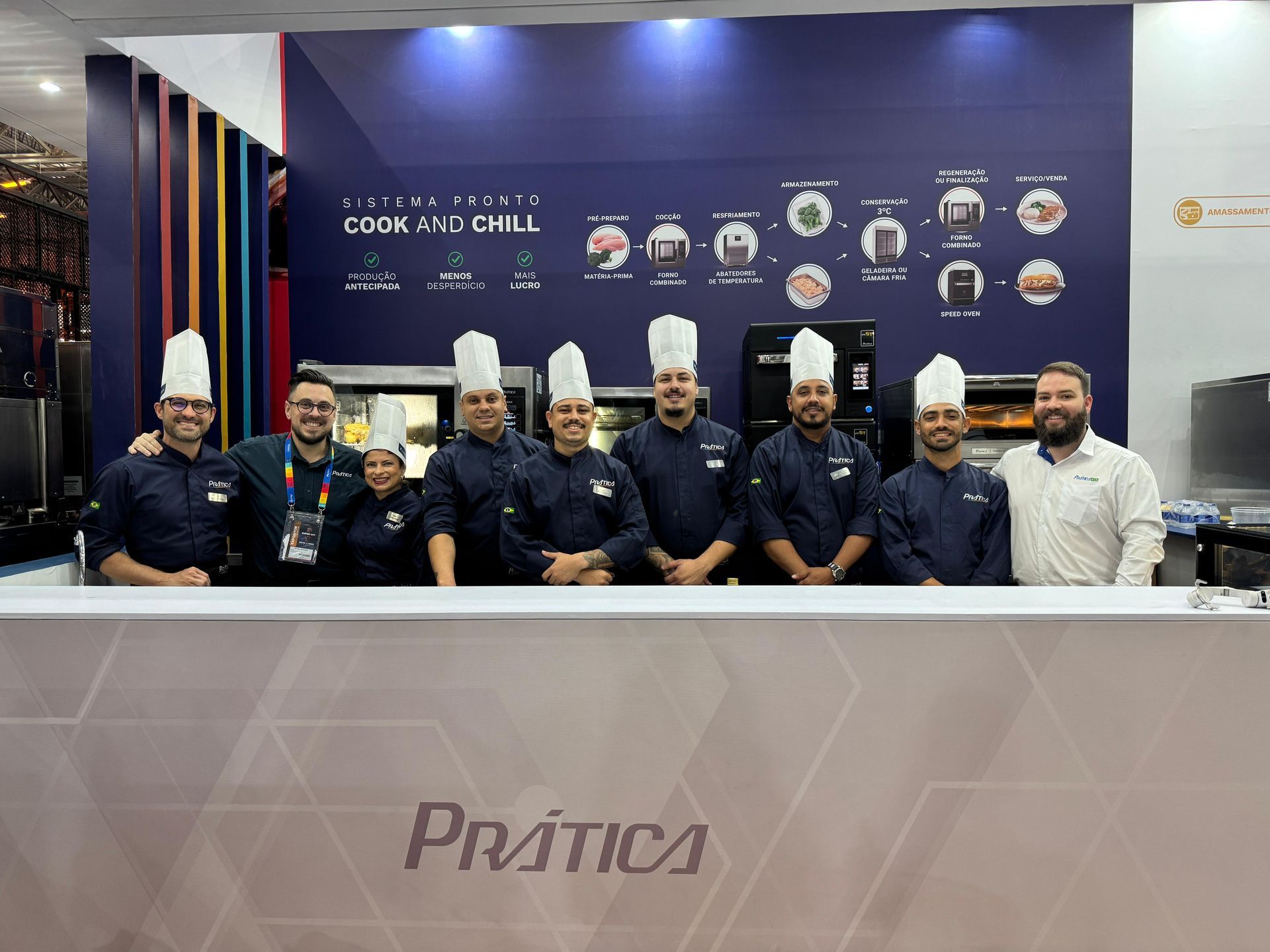 Chefs com chapéus brancos e uniformes azuis posam atrás de um balcão com o logotipo 