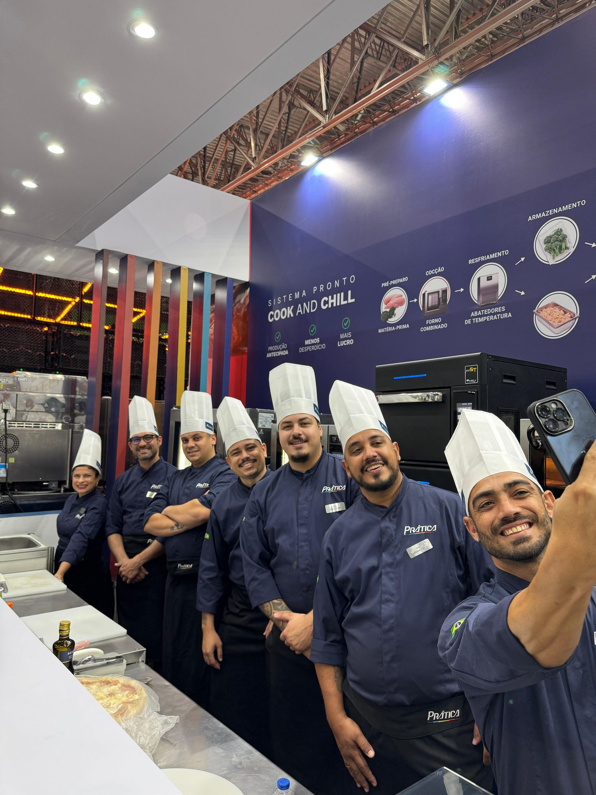 Chefs com uniformes azuis e chapéus brancos tiram uma selfie em uma cozinha com diversos utensílios.