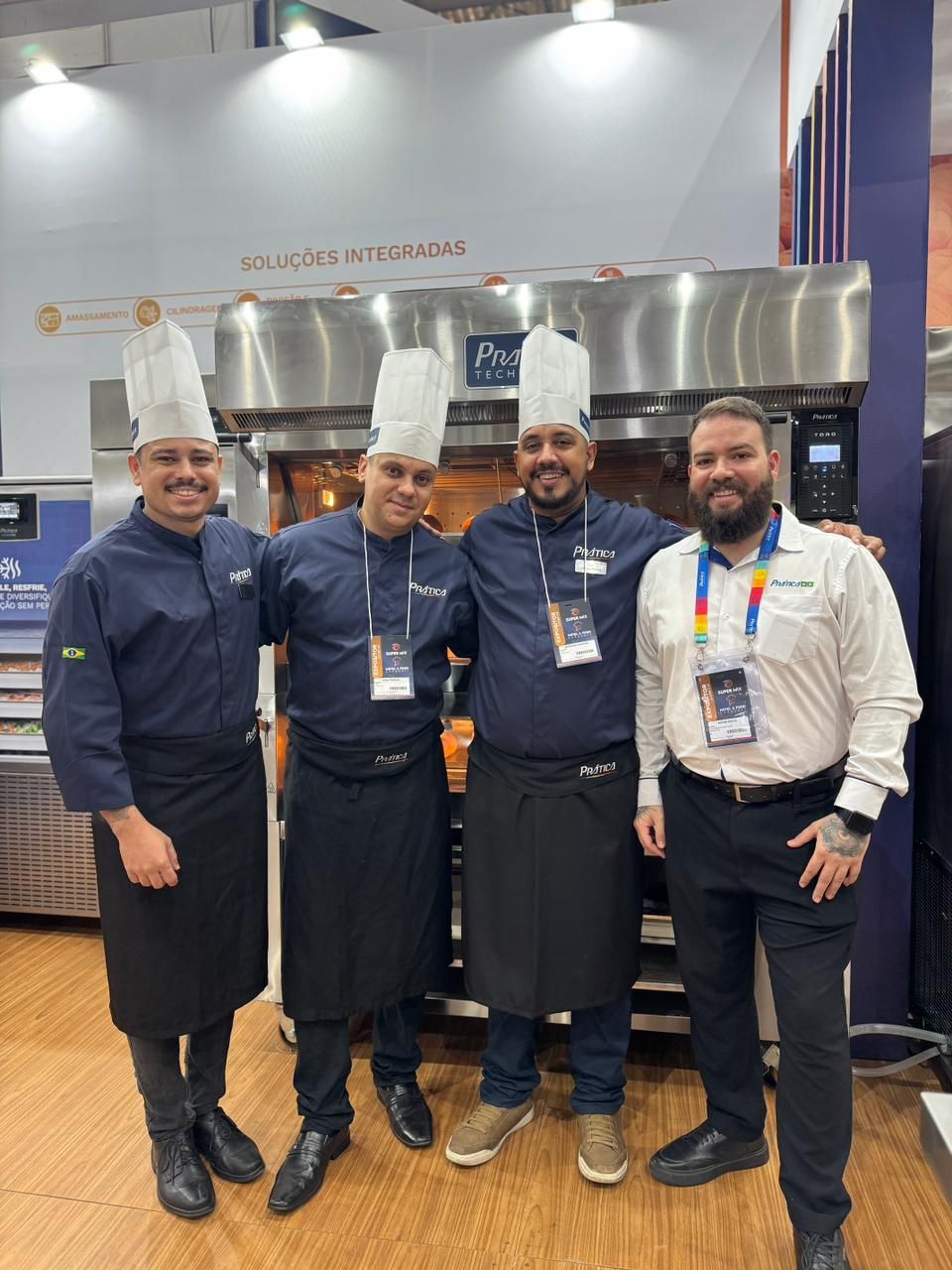 Quatro homens em frente a um forno industrial. Três usam chapéus e uniformes de chef, posando com o forno. O quarto homem veste uma camisa branca.