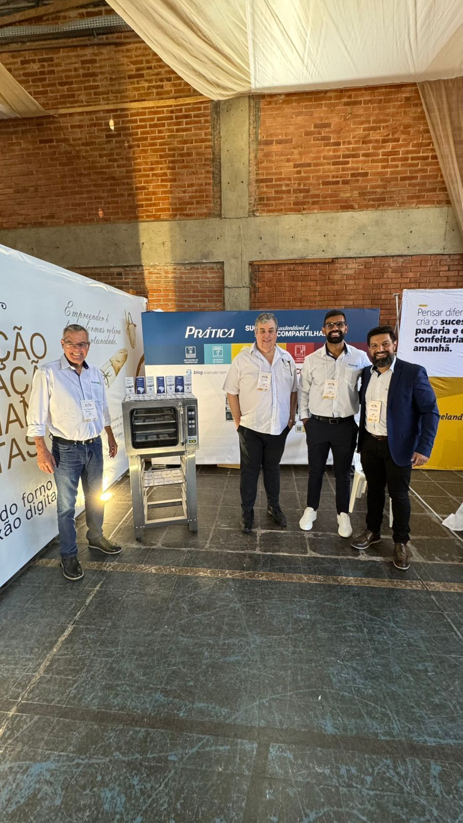 Quatro homens em pé ao lado de um forno industrial em um espaço bem iluminado, com faixas e uma parede de tijolos.