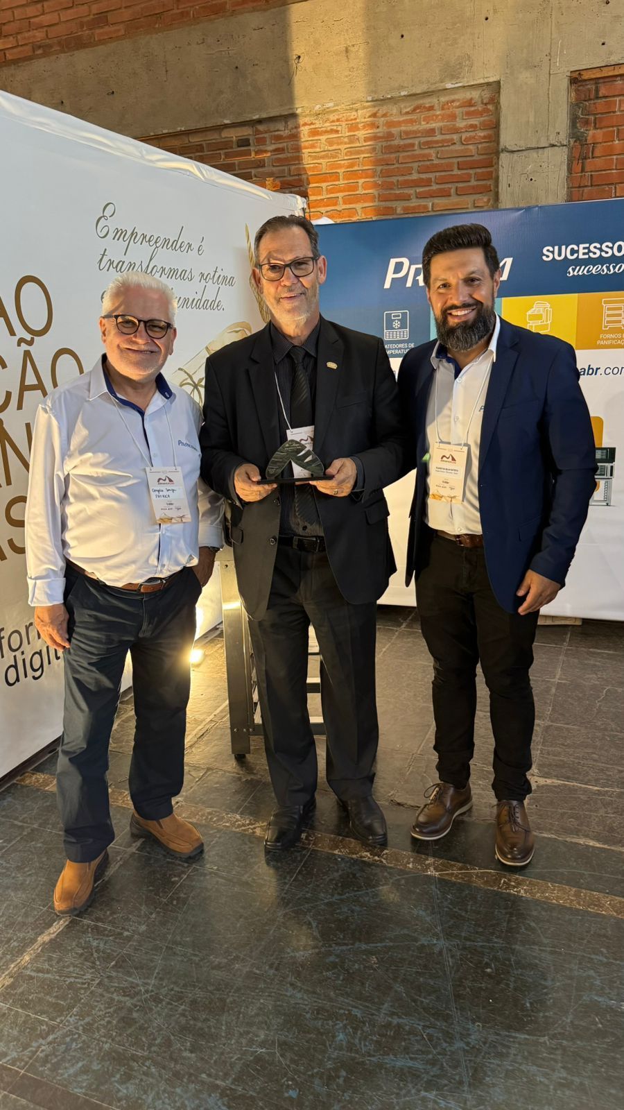 Três homens em trajes de negócios posam para uma foto. Um deles segura um troféu. Cenário de evento.