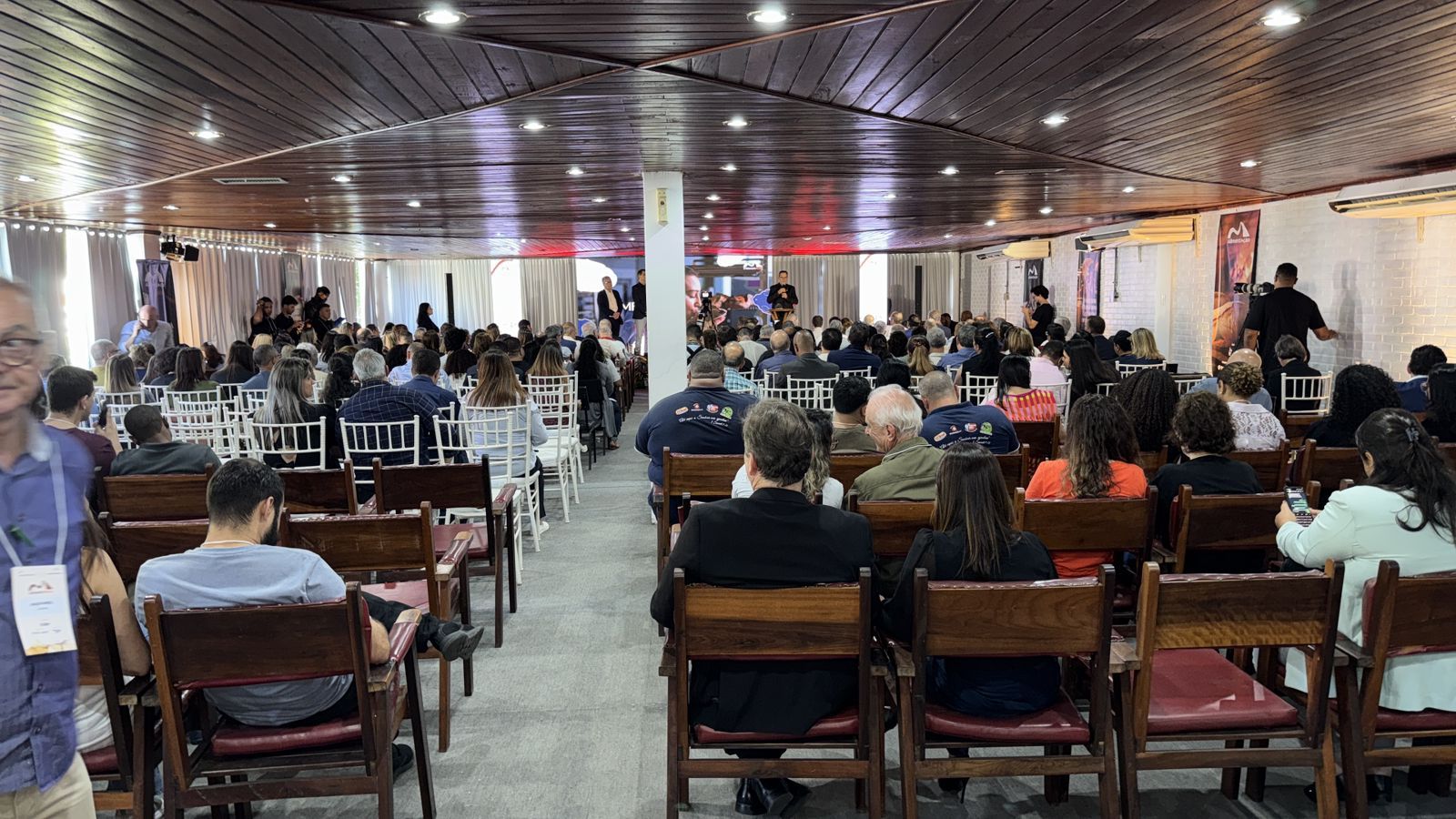 Grande salão de conferências com fileiras de pessoas sentadas de frente para um palco, paredes brancas e cadeiras de madeira.