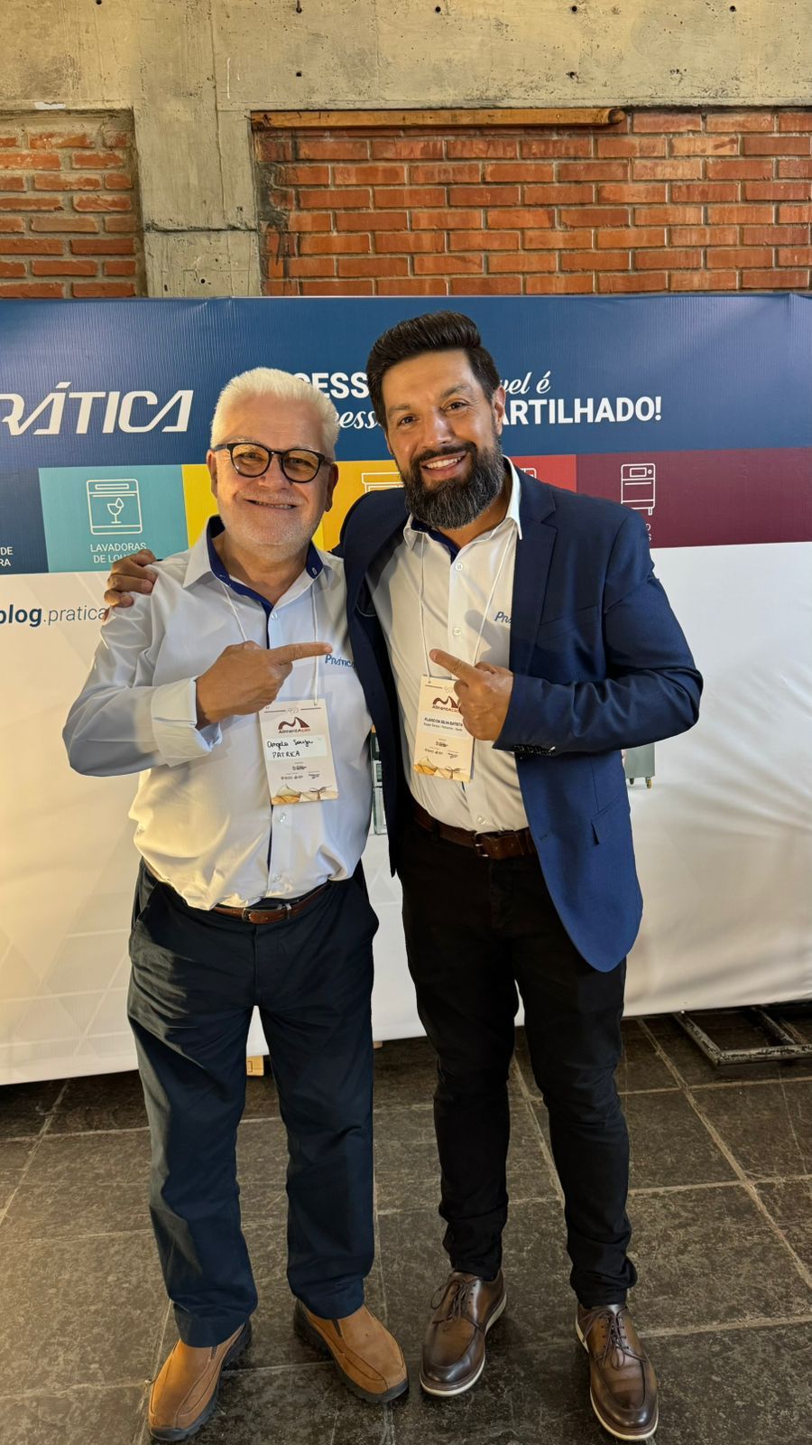 Dois homens sorrindo e apontando. Um de camisa azul-clara, o outro de blazer. Estão em pé perto de uma faixa da conferência.