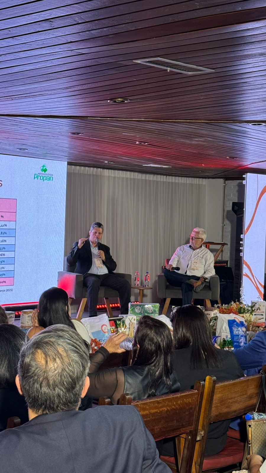Dois homens no palco de uma conferência, falando para uma plateia.