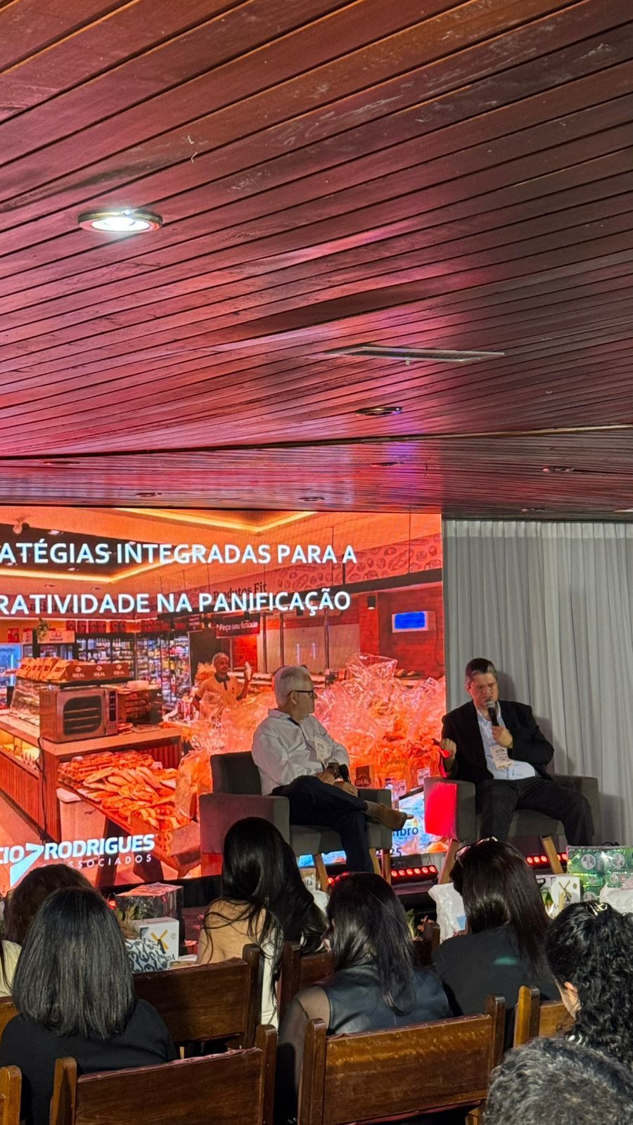 Dois homens no palco, discursando em um evento; plateia sentada à sua frente; tela ao fundo.
