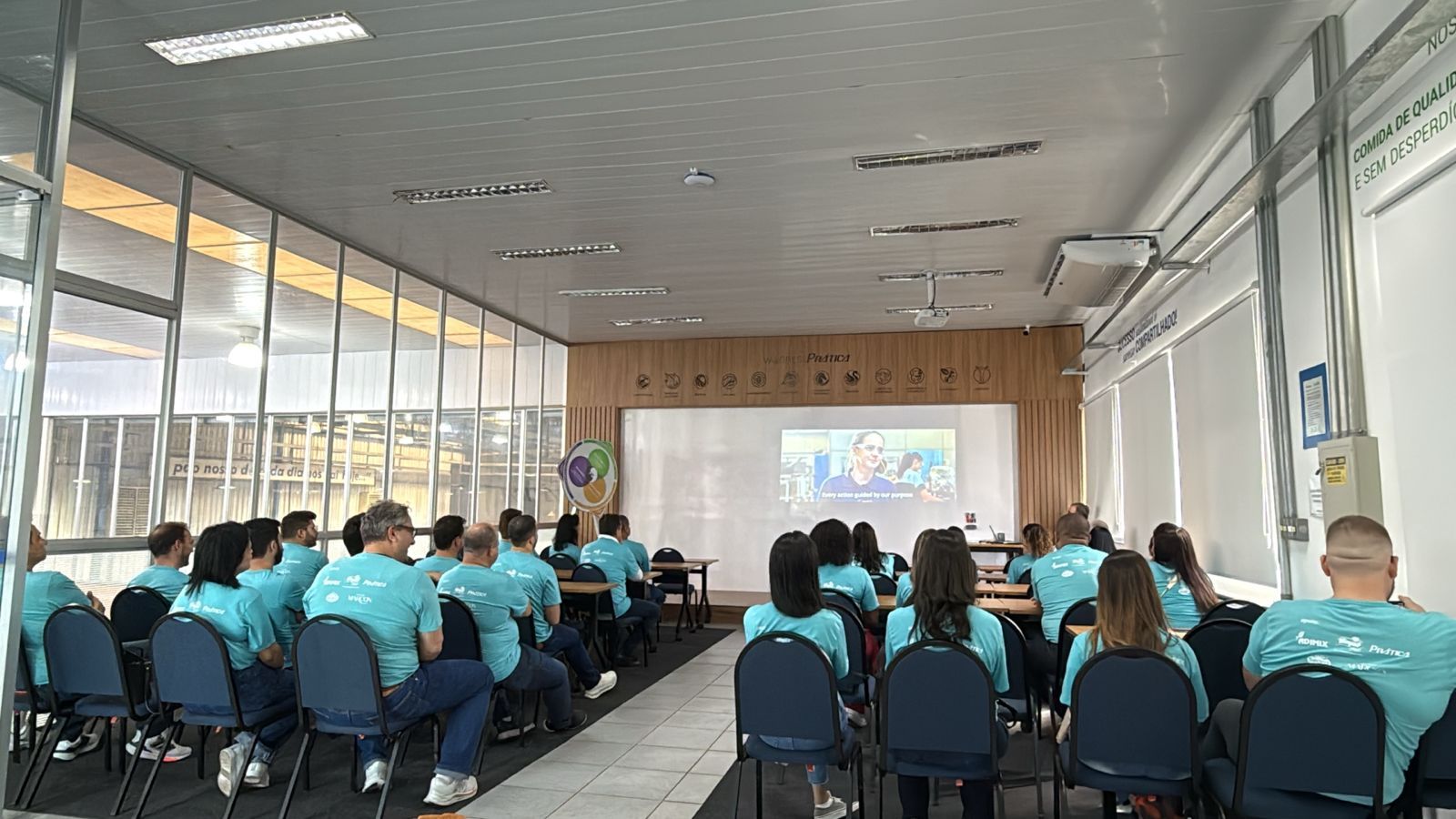 Pessoas vestindo camisetas verde-azuladas sentadas em uma sala de aula, assistindo a uma apresentação em uma tela.