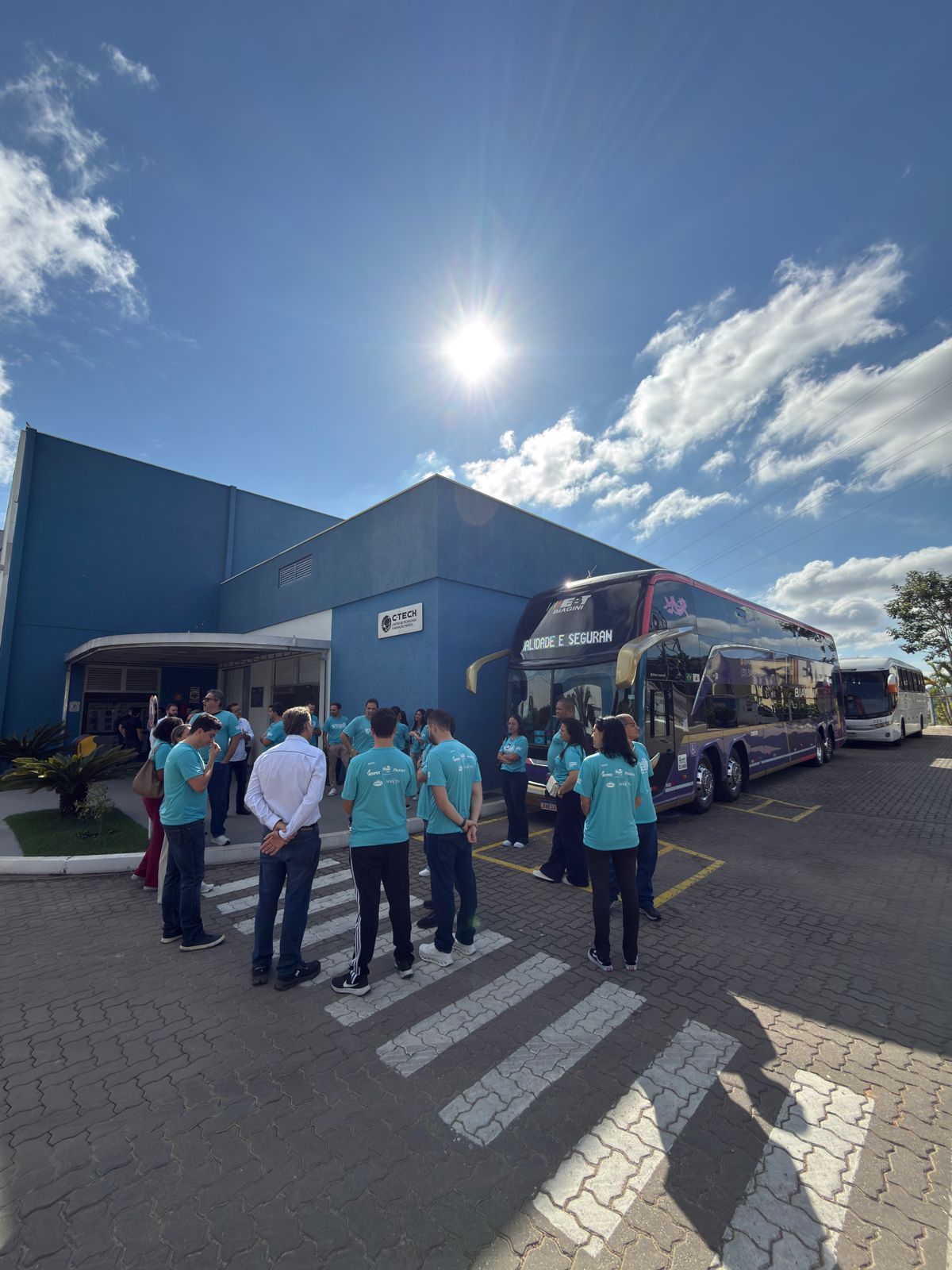 Pessoas vestindo camisetas verde-azuladas estão perto de ônibus em frente a um prédio azul sob um céu ensolarado.