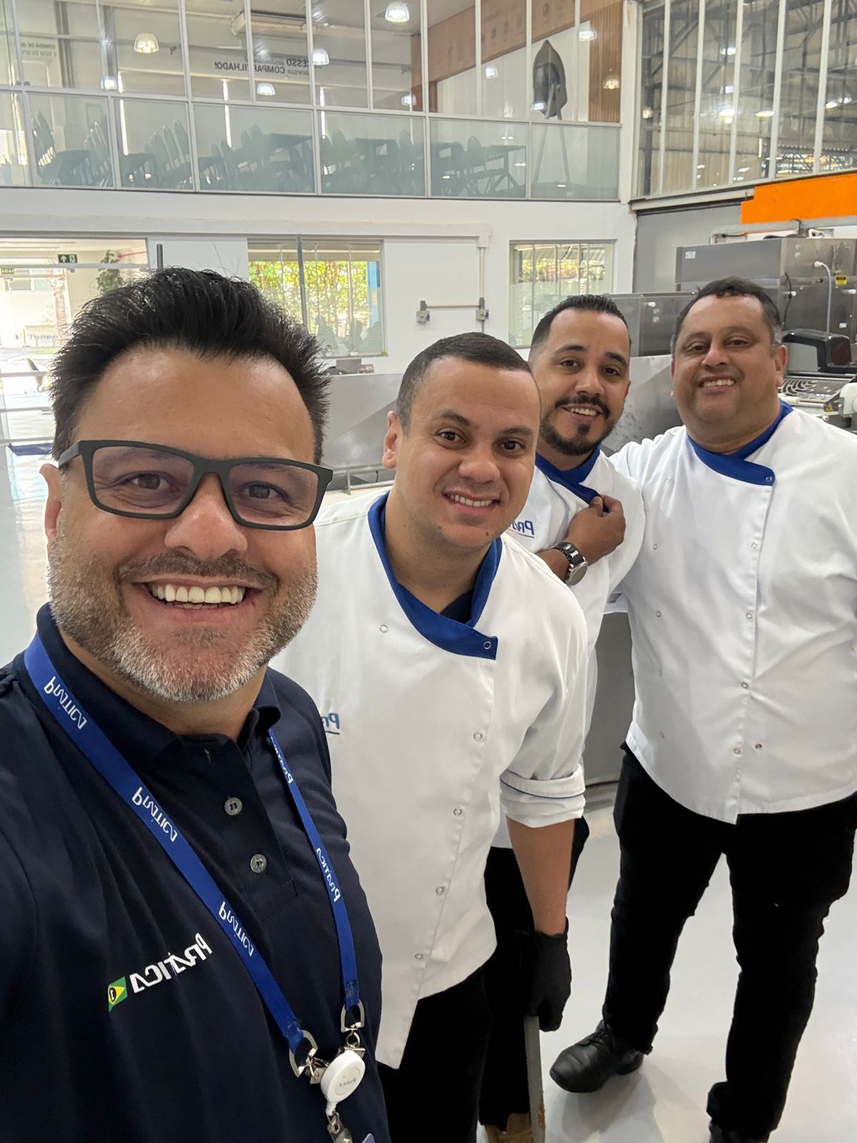 Quatro homens de avental de chef e um homem de óculos sorriem para uma selfie em uma cozinha.