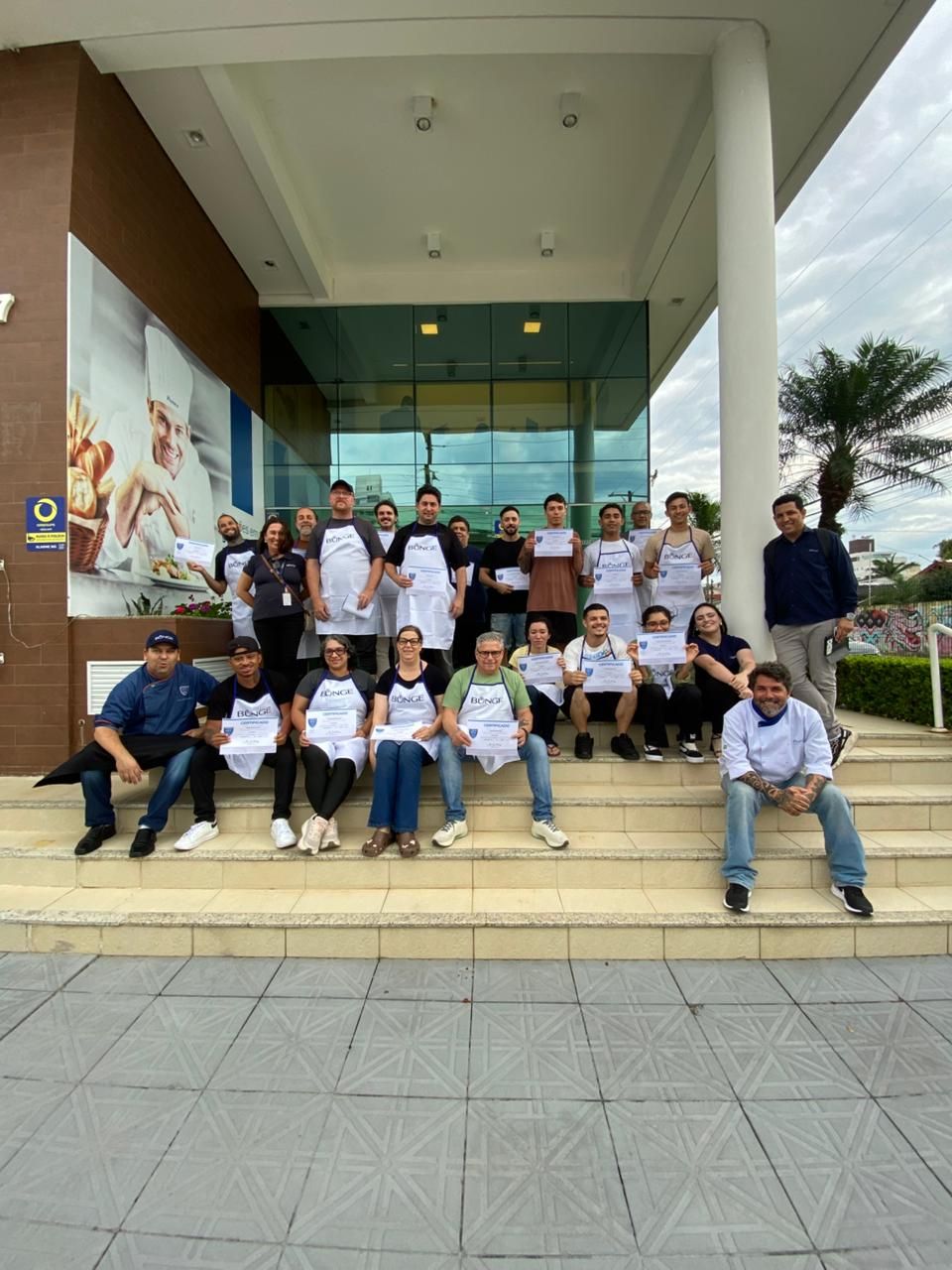Grupo de pessoas de avental segurando certificados em frente a um prédio com portas de vidro e uma placa.