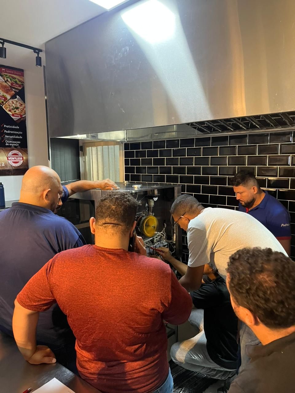 Cinco homens estão reunidos em torno de equipamentos de cozinha, provavelmente fazendo reparos, embaixo de uma saída de ventilação de aço inoxidável.