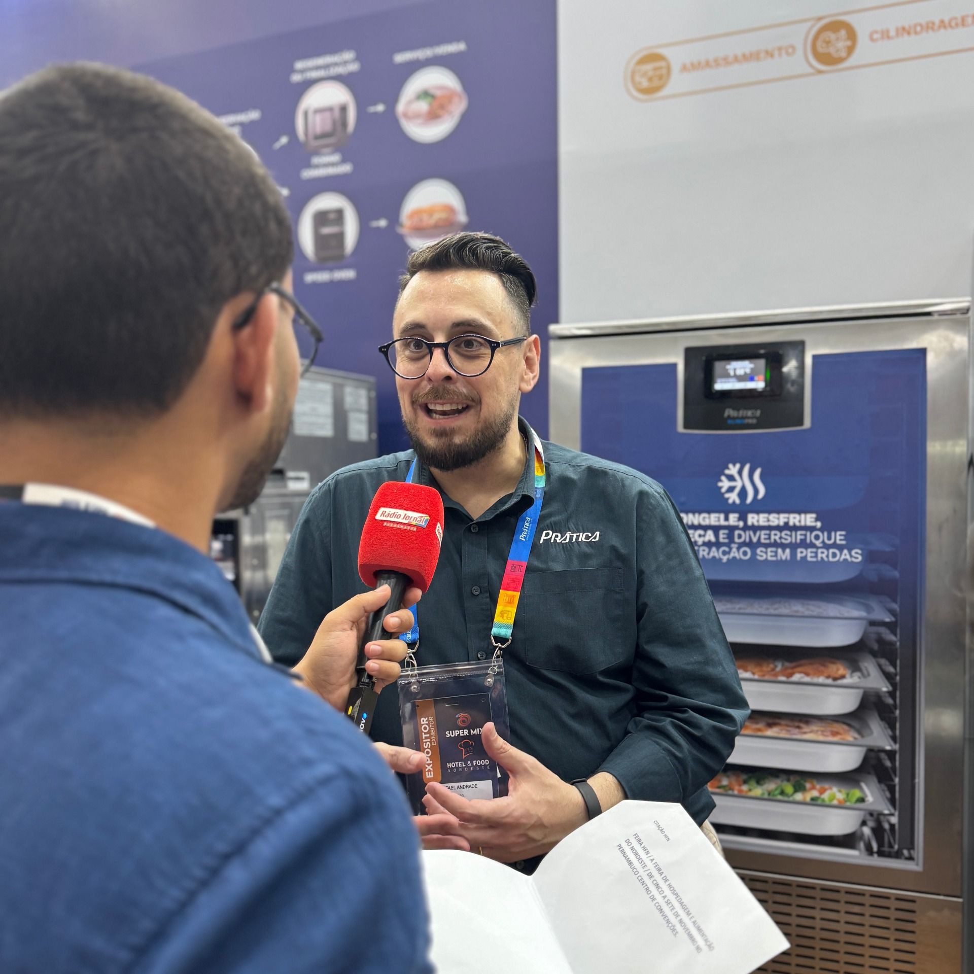 Homem sendo entrevistado em frente a um freezer comercial. Ele segura um recipiente transparente e sorri.
