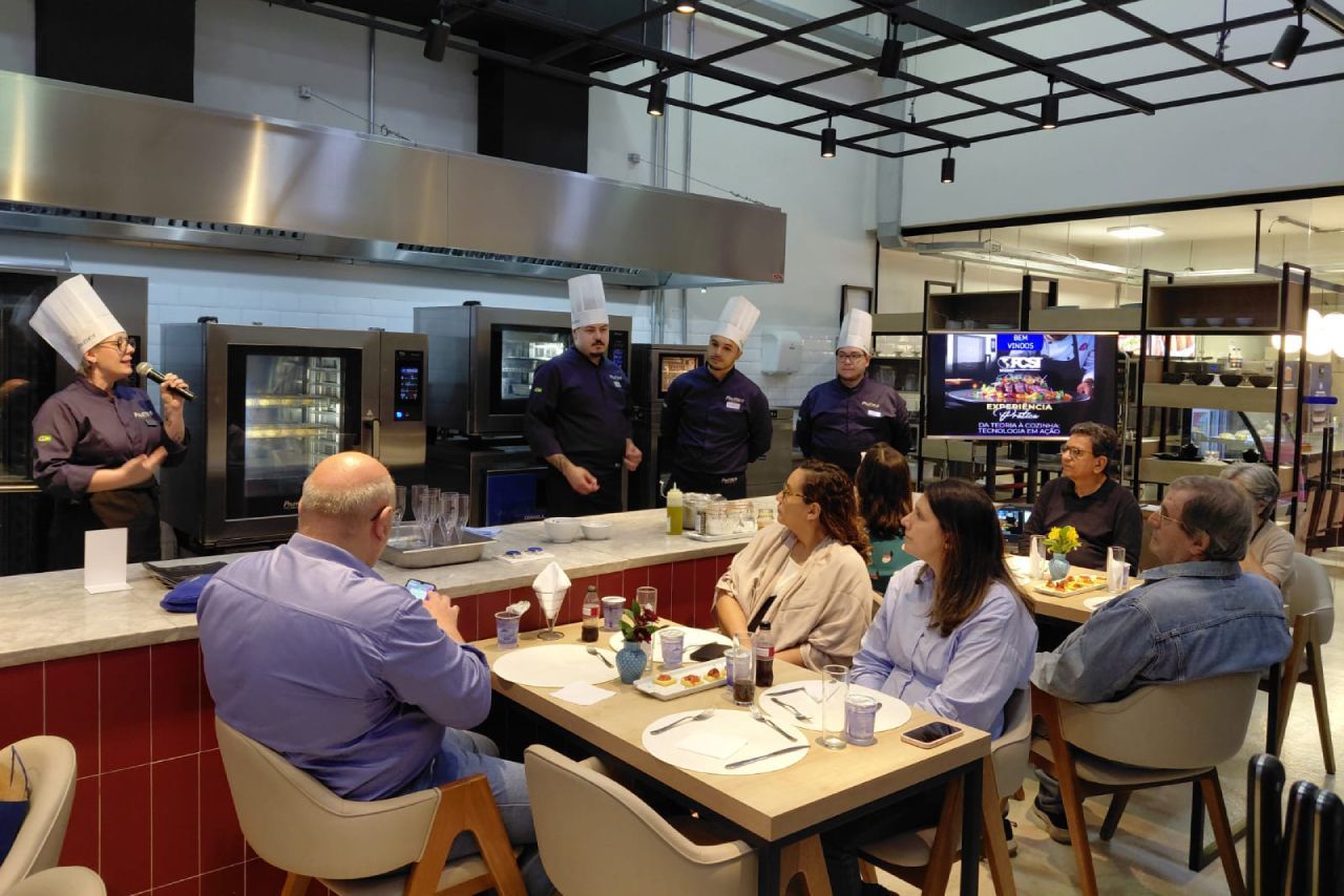 Uma chef de uniforme branco falando, vários chefs atrás dela. Convidados sentados às mesas, ambiente de cozinha industrial.