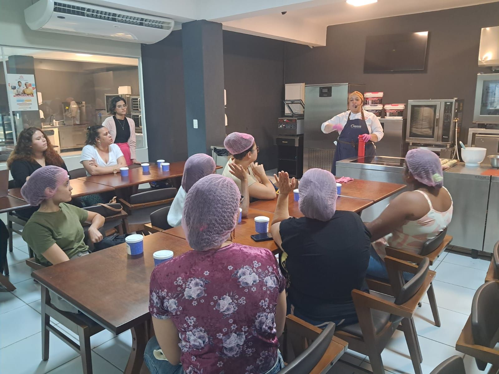 Uma aula de culinária em uma cozinha. Instrutor gesticula enquanto mulheres com redes nos cabelos escutam nas mesas.