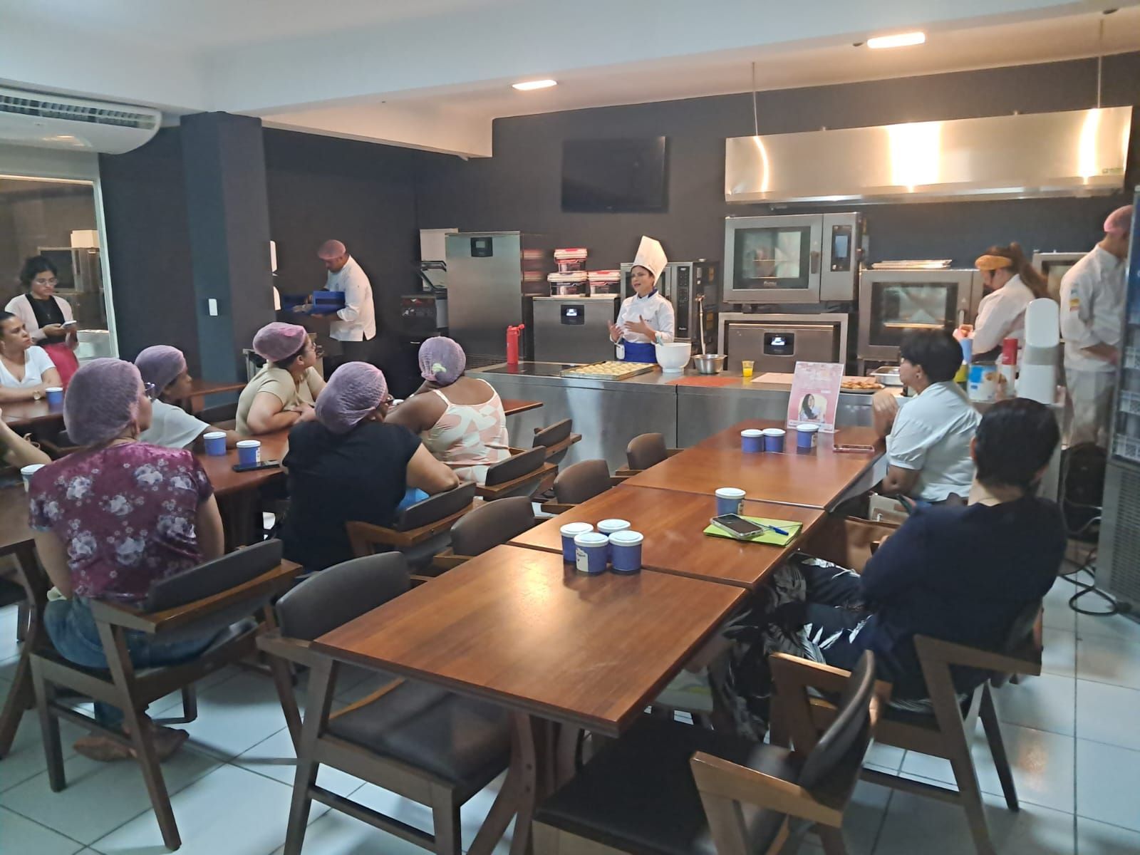 Uma aula de culinária em uma cozinha. Participantes com toucas assistem a uma demonstração de um chef.