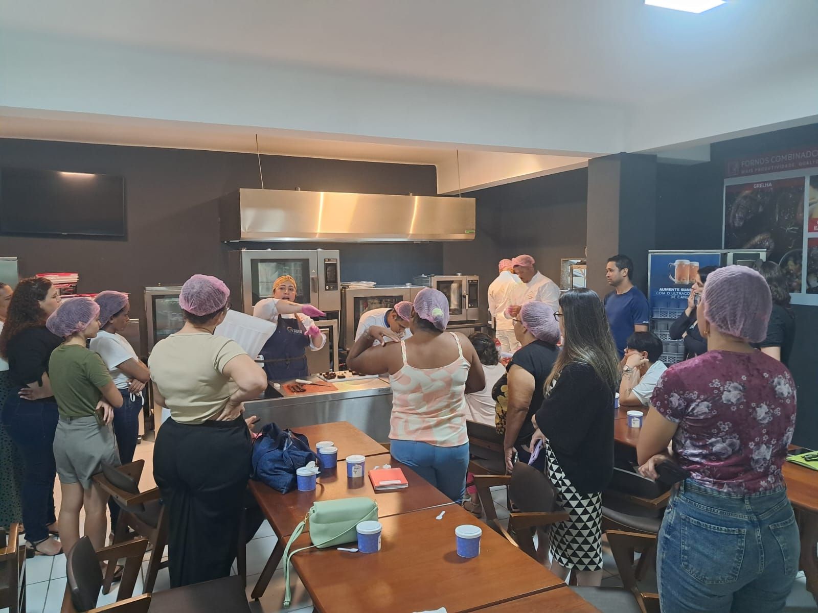 Uma aula de culinária em uma cozinha. Pessoas usando toucas assistem à demonstração de um chef.