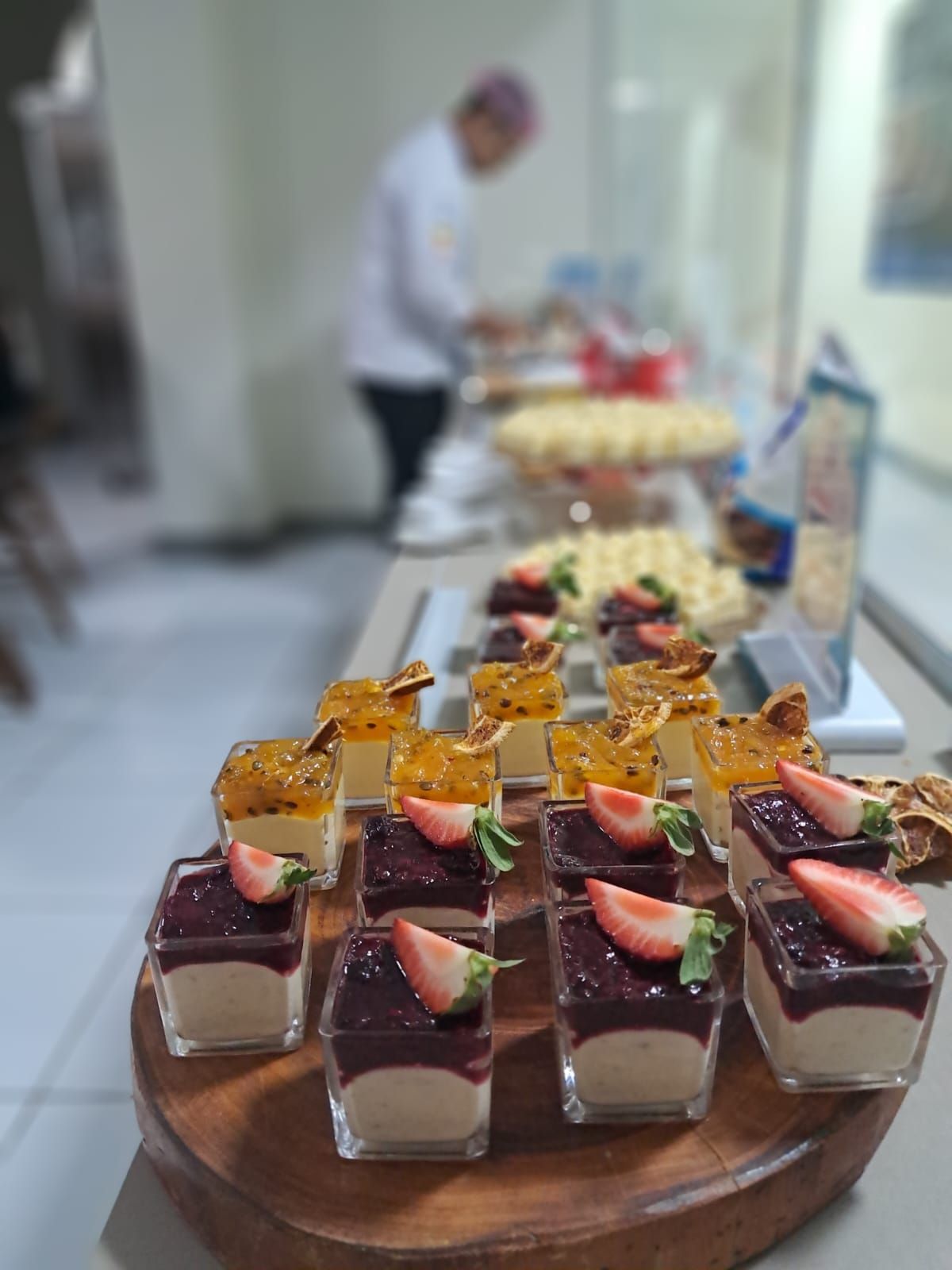 Buffet de sobremesas com copos quadrados de vidro com mousse coberta com frutas, em frente a um chef.
