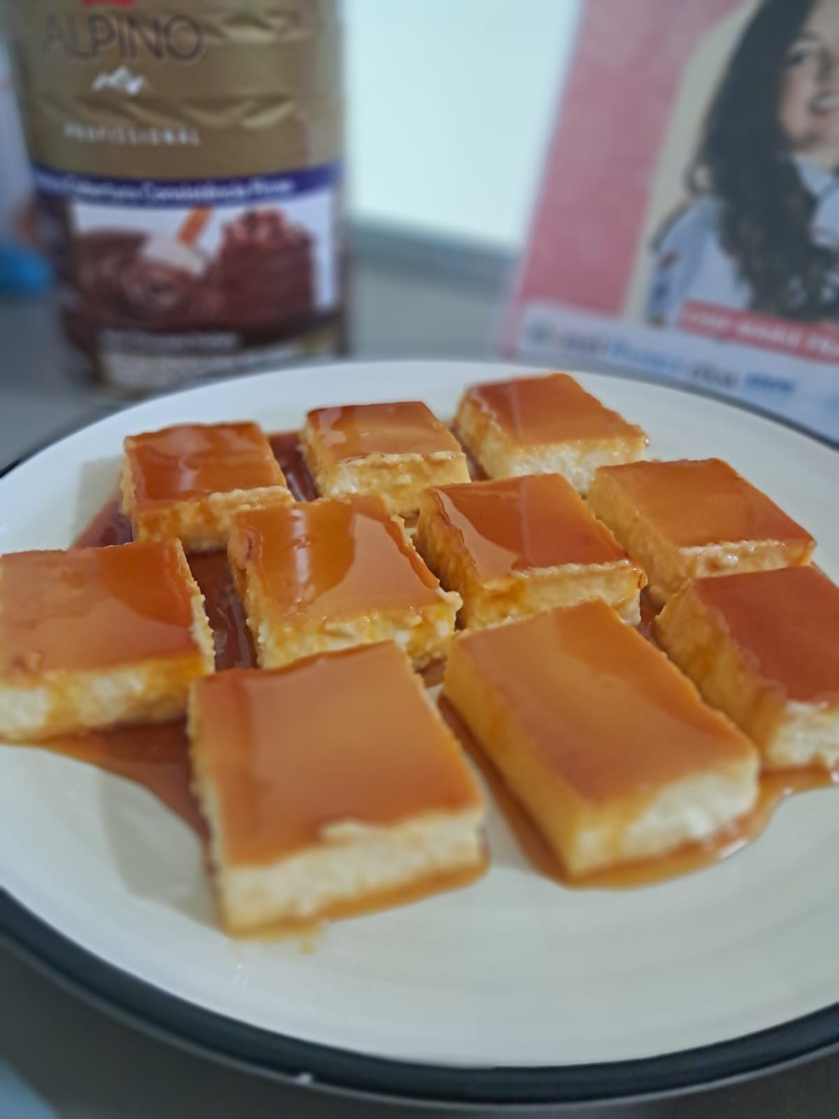 Um prato de quadrados de creme de caramelo com uma parte superior brilhante, ao lado de um pote e um folheto.