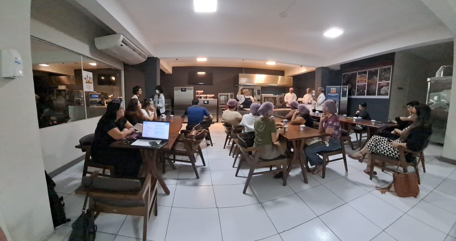 Um grupo de pessoas se reúne em um café, sentadas em mesas e cadeiras, possivelmente participando de uma reunião ou evento.