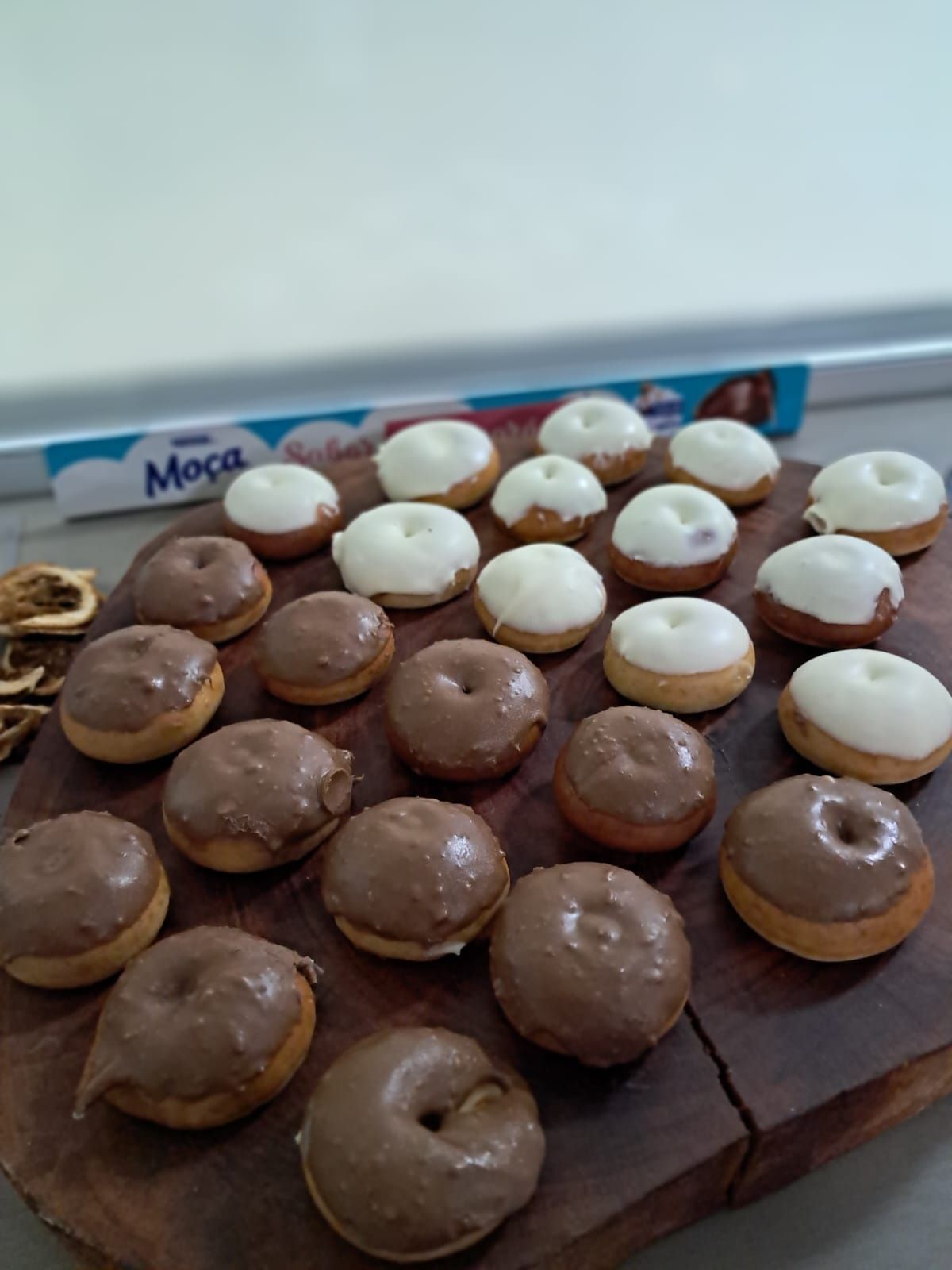 Mini donuts, alguns com cobertura de chocolate, outros com cobertura branca, dispostos sobre uma tábua de madeira. Uma caixa é parcialmente visível ao fundo.