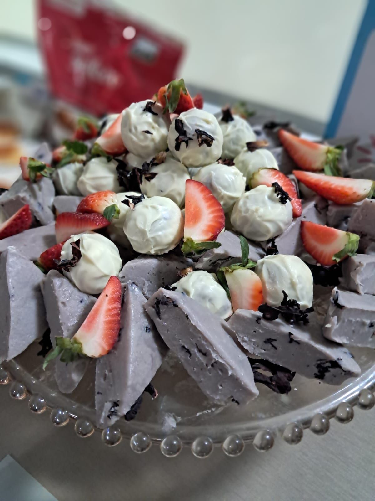 Prato de sobremesa com trufas de chocolate branco, quadrados roxos e morangos fatiados, apresentados em um suporte de vidro para bolos.