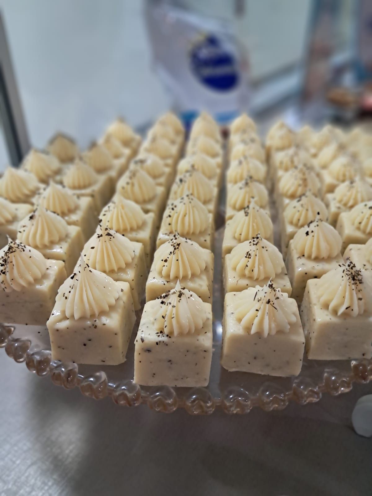 Fileiras de pequenos pastéis quadrados com cobertura de creme e sementes de papoula em uma bandeja de vidro.
