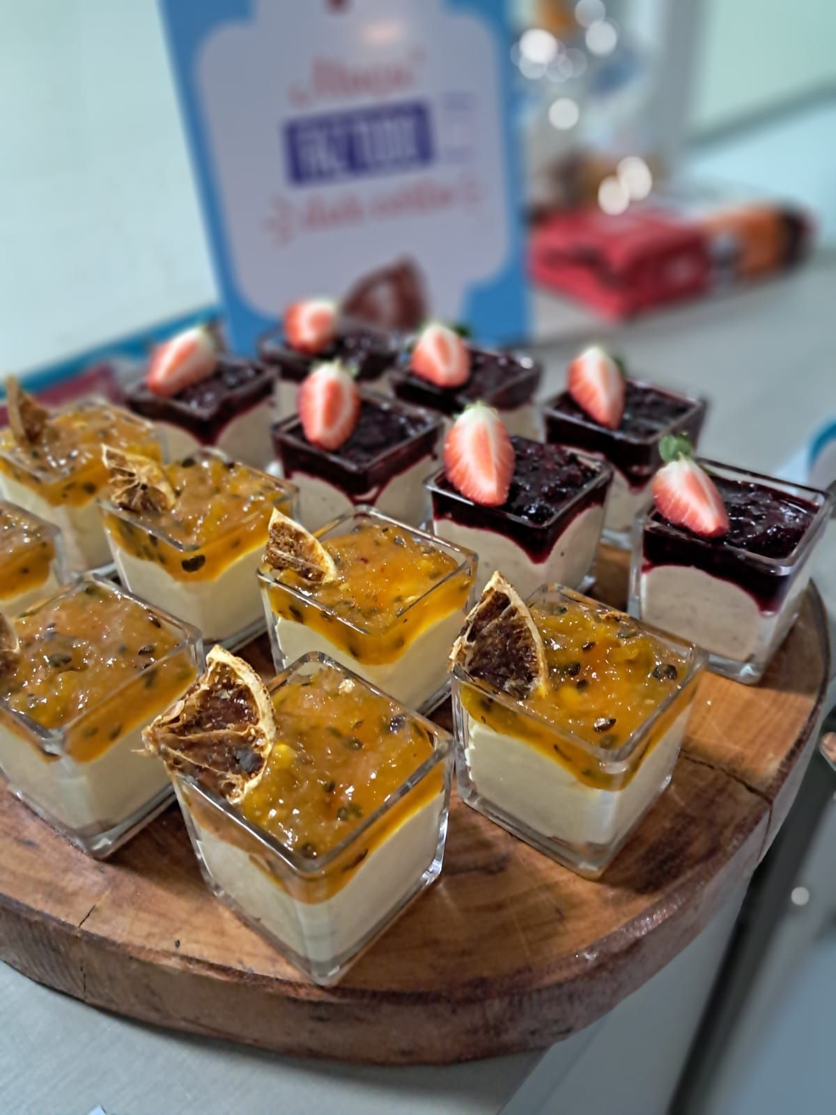 Mini cheesecakes com cobertura de maracujá e frutas vermelhas, dispostos sobre uma tábua de madeira, com uma placa desfocada ao fundo.