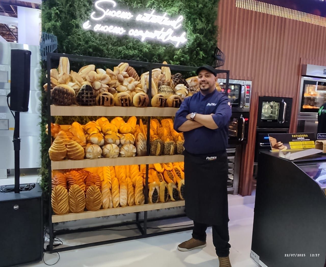 Padeiro orgulhoso em frente a uma vitrine com diversos pães. Parede verde, placa e cenário de cozinha comercial.